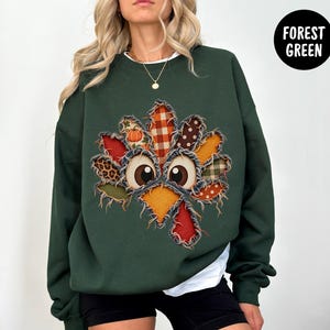 May include: A forest green crewneck sweatshirt featuring a central turkey graphic composed of various fabric patches with frayed edges. The turkey design includes patterns like plaid, polka dots, and leopard print, with large cartoon eyes.