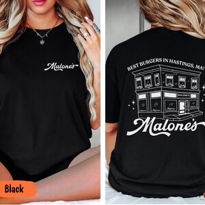 May include: A black t-shirt featuring the white text Malone's on the front chest. The back displays a white line drawing of a building with the text Best Burgers in Hastings, MA! and the word Malone's in a large script font below the building.