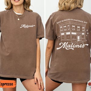 May include: A brown short-sleeve t-shirt featuring the white text Malone's on the front left chest. The back displays a white line drawing of a building with the text Best Burgers In Hastings, MA! and the word Malone's in a script font.
