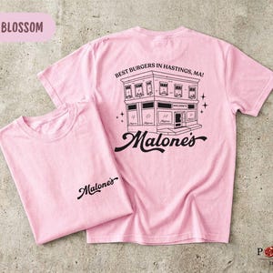 May include: A light pink short-sleeve t-shirt featuring a black graphic of a building with the text "BEST BURGERS IN HASTINGS, MA!" and "Malone's" on the back, and the word "Malone's" printed in black on the front left chest area.