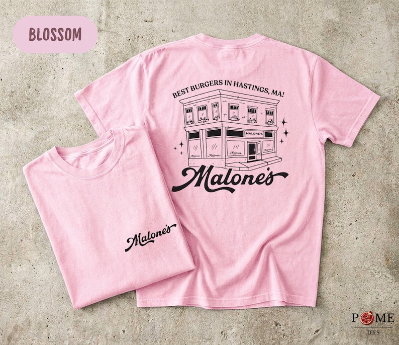 May include: A light pink short-sleeve t-shirt featuring a black graphic of a building with the text "BEST BURGERS IN HASTINGS, MA!" and "Malone's" on the back, and the word "Malone's" printed in black on the front left chest area.
