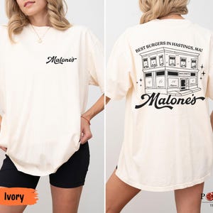 May include: An ivory t-shirt featuring a small black script logo on the front chest that reads Malone's. The back displays a large black graphic of a building with the text Best Burgers in Hastings, MA! and the script logo Malone's below it.