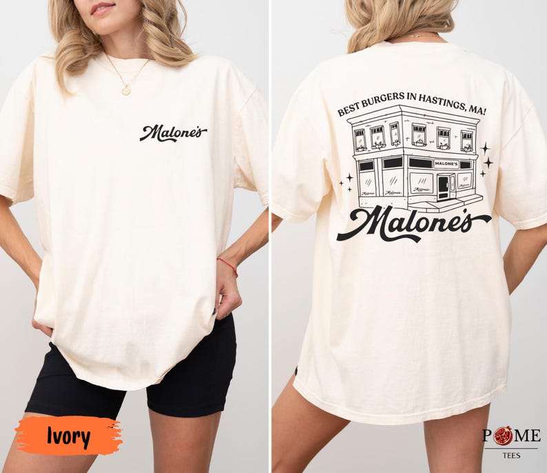 May include: An ivory t-shirt featuring a small black script logo on the front chest that reads Malone's. The back displays a large black graphic of a building with the text Best Burgers in Hastings, MA! and the script logo Malone's below it.