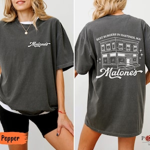 May include: A charcoal gray short-sleeve t-shirt featuring a white script logo on the front chest. The back displays a white line-art illustration of a building with the text "BEST BURGERS IN HASTINGS, MA!" and the script logo "Malone's" below.
