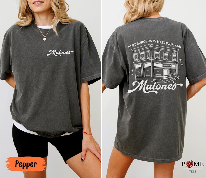 May include: A charcoal gray short-sleeve t-shirt featuring a white script logo on the front chest. The back displays a white line-art illustration of a building with the text "BEST BURGERS IN HASTINGS, MA!" and the script logo "Malone's" below.