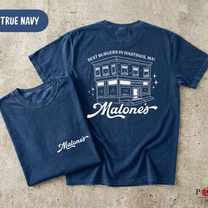 May include: A true navy blue short-sleeve t-shirt featuring a white graphic print. The front displays the text Malone's. The back features a detailed line drawing of a building with the text Best Burgers In Hastings, MA! and Malone's.