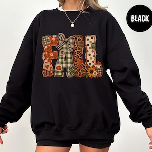 May include: A black long-sleeve sweatshirt featuring the word FALL in large, block-style letters. Each letter is filled with a different patchwork pattern, including orange floral, green plaid, leopard print, and polka dots, accented with a bow.