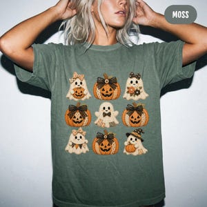 May include: A moss green short-sleeve t-shirt featuring a graphic print of nine individual icons arranged in a grid. The icons include various ghosts and pumpkins with patterned textures, bows, and hats. The word MOSS is printed in the top corner.