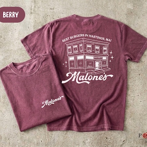 May include: A berry-colored short-sleeve t-shirt featuring a white graphic print. The front displays the text 'Malone's' in script. The back shows a building illustration with the text 'BEST BURGERS IN HASTINGS, MA!' and 'Malone's' in script.