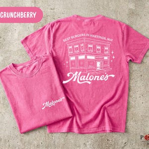 May include: A bright pink short-sleeve t-shirt featuring a white graphic on the back of a building with the text 'BEST BURGERS IN HASTINGS, MA!' and 'Malone's'. The front chest area displays the white script text 'Malone's' in the upper left corner.