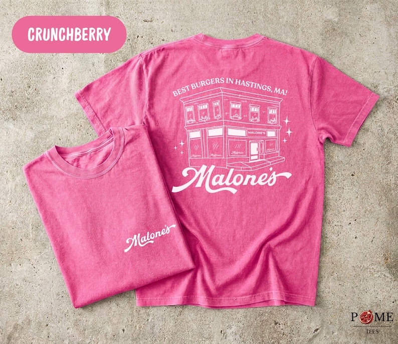 May include: A bright pink short-sleeve t-shirt featuring a white graphic on the back of a building with the text 'BEST BURGERS IN HASTINGS, MA!' and 'Malone's'. The front chest area displays the white script text 'Malone's' in the upper left corner.