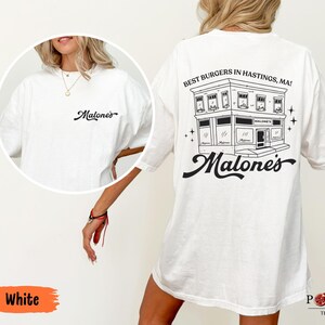 May include: A white t-shirt featuring a black graphic on the back of a building with the text "BEST BURGERS IN HASTINGS, MA!" and "Malone's" in a script font. The front chest area displays the same "Malone's" script logo in black ink.