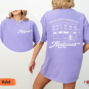 May include: A violet short-sleeve t-shirt featuring white screen-printed graphics. The front displays the text "Malone's" in script. The back features a line-art illustration of a building with the text "BEST BURGERS IN HASTINGS, MA!" and "Malone's".