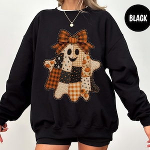 May include: A black long-sleeve sweatshirt featuring a central graphic of a ghost made of various patchwork patterns in orange, black, and white. The ghost has a smiling face, a large plaid bow on its head, and small pumpkin illustrations.