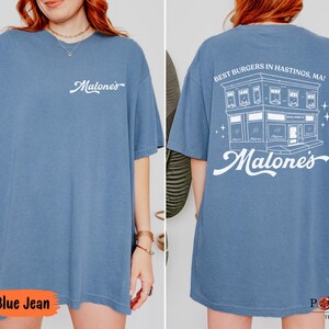 May include: A blue jean colored t-shirt featuring a white script logo on the front chest. The back displays a white graphic of a building with the text "BEST BURGERS IN HASTINGS, MA!" and "Malone's" in a large script font below the building.