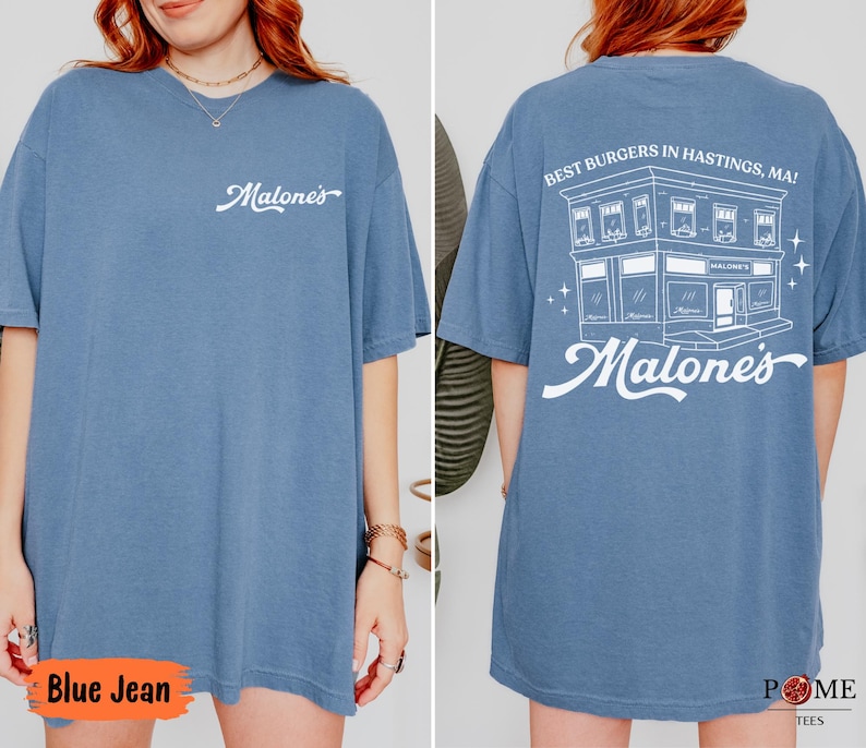 May include: A blue jean colored t-shirt featuring a white script logo on the front chest. The back displays a white graphic of a building with the text "BEST BURGERS IN HASTINGS, MA!" and "Malone's" in a large script font below the building.