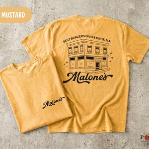 May include: Mustard yellow short-sleeve t-shirt featuring a black graphic of a building with the text "BEST BURGERS IN HASTINGS, MA!" and "Malone's" on the back, and a smaller "Malone's" logo printed on the front chest area of the folded shirt.