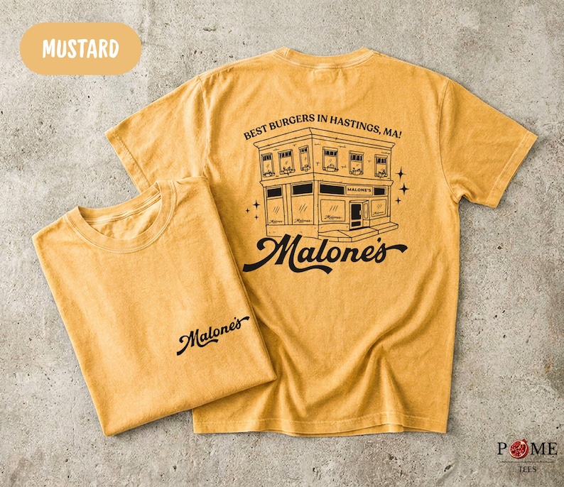 May include: Mustard yellow short-sleeve t-shirt featuring a black graphic of a building with the text "BEST BURGERS IN HASTINGS, MA!" and "Malone's" on the back, and a smaller "Malone's" logo printed on the front chest area of the folded shirt.