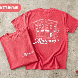 May include: A watermelon red short-sleeve t-shirt featuring a white graphic on the back of a building with the text BEST BURGERS IN HASTINGS, MA! and Malone's. The front left chest area displays the white text Malone's in a cursive script font.