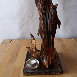 Könnte beinhalten: Eine rustikale Holzskulptur mit natürlicher, organischer Form. Das dunkelbraune Holz hat eine strukturierte Oberfläche und ist auf einem quadratischen Sockel montiert. Ein kleiner, klarer Glaskerzenhalter mit einer brennenden Teelichtkerze befindet sich auf dem Sockel.