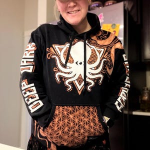 May include: Black hoodie with an octopus design in white and orange. The hoodie has a kangaroo pocket with an orange geometric pattern. The sleeves have the words "DEEP DARK" printed in white. A camouflage baseball cap is worn.