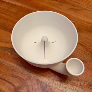 May include: A white, circular kitchen gadget with a central post and a spout. The gadget has a bowl-like shape with a small, cylindrical spout extending from the side. The design is minimalist and modern, suitable for food preparation.