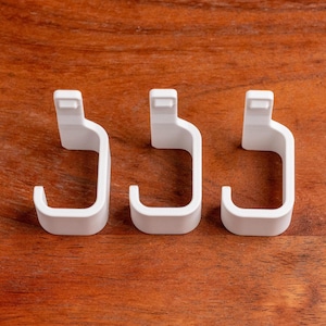 May include: Three white, J-shaped hooks are arranged on a wooden surface. The hooks have a rectangular top and a curved body with a hook at the bottom. The background is a warm, brown wood grain.