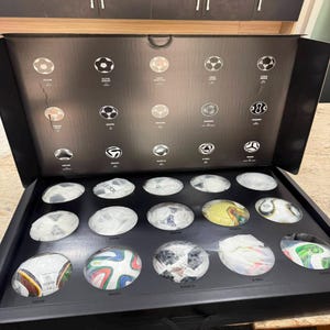 以下が含まれることがあります： ミニチュアサッカーボールのコレクションが入った黒いプレゼンテーションボックス。内蓋には様々なサッカーボールのデザインの白い線画とテキストラベルが描かれており、ベース部分には15個の個別のサッカーボールが収められています。