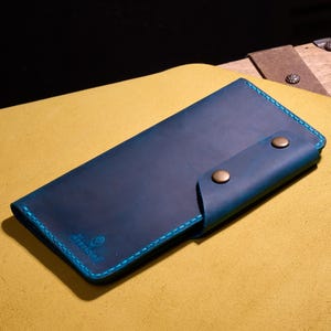 Op de afbeelding: Een handgemaakte, blauwe leren portemonnee met contrasterende turquoise stiksels en afwerking. De portemonnee heeft een klepsluiting die wordt vastgezet met twee messingkleurige drukknopen. De merknaam is in de linkerbenedenhoek in reliëf aangebracht.