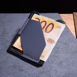 Może przedstawiać: Minimalistyczny portfel z czarnej i szarej skóry zawiera stos banknotów. Portfel ma geometryczny kształt z szarym panelem przednim i jest wypełniony walutą. Tło to teksturowana szara powierzchnia.