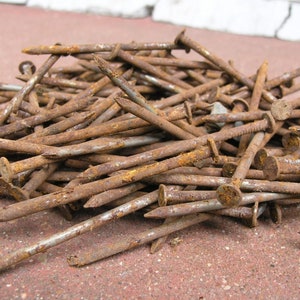 May include: A pile of rusty nails with flat, circular heads and pointed tips. The nails are various lengths and show signs of corrosion, with a reddish-brown color. They are scattered on a reddish-brown surface.