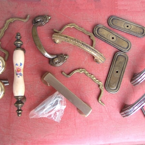 May include: A collection of vintage cabinet hardware, including handles, pulls, and backplates. The hardware features various designs, including floral patterns, brass, and chrome finishes. Some pieces have decorative details and are arranged on a red surface.