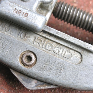 Puede incluir: Primer plano de un cortatubos Ridgid vintage, modelo No. 10. La herramienta est&aacute; hecha de metal gris desgastado con texto visible y un tornillo de ajuste roscado. El cortador muestra signos de desgaste.