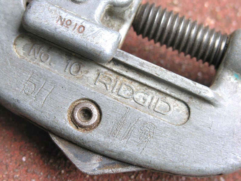Puede incluir: Primer plano de un cortatubos Ridgid vintage, modelo No. 10. La herramienta est&aacute; hecha de metal gris desgastado con texto visible y un tornillo de ajuste roscado. El cortador muestra signos de desgaste.
