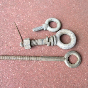 May include: Three silver-toned metal eye bolts of varying sizes. The top bolt is short with a threaded shank. The middle bolt features a hexagonal nut and a thin metal pin. The bottom bolt is long with a fully threaded shank and a circular eye.