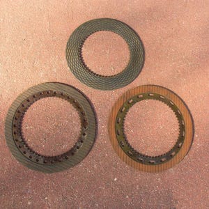 May include: Three circular mechanical parts with textured surfaces. The top part is dark gray with a diamond pattern. The bottom left part is gray with a ridged pattern and small holes. The bottom right part is brown with a ridged pattern and a green inner ring.