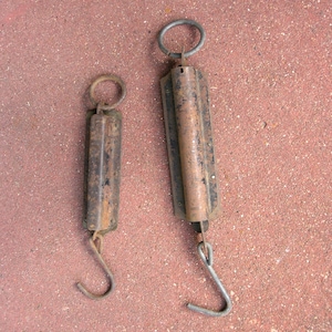 May include: Two vintage, rusty metal hanging scales. One is smaller than the other. Each scale has a metal ring at the top for hanging, a rectangular body, and a hook at the bottom. The scales are likely used for weighing items.