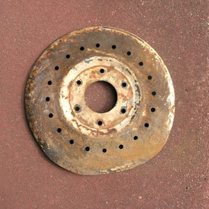 May include: A rusty, circular metal object with a central hole and numerous smaller holes around the perimeter. The object has a weathered appearance with shades of brown, white, and black, suggesting age and exposure to the elements. The object is likely a brake rotor.