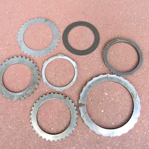 May include: An overhead shot displays a collection of circular metal components. The pieces vary in size and design, with some featuring toothed edges and others smooth rims. The components are arranged on a reddish-brown surface, suggesting an industrial or mechanical context.