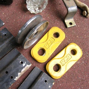 May include: Assorted hardware components including yellow, metal pieces with the letters "RCS", metal brackets, and a clear glass knob. The items are arranged on a textured, reddish-brown surface.
