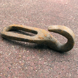 May include: A weathered, rusty metal hook with an oval eye and a curved hook end. The hook lies on a textured, reddish-brown surface. The metal shows signs of age and exposure to the elements.