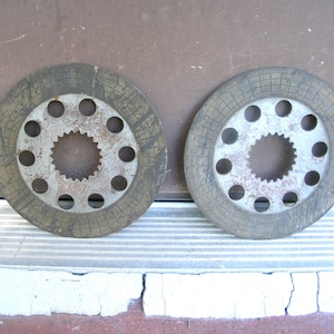 May include: Two used, circular brake rotors. Each rotor has a central, toothed hub surrounded by a ring of holes. The outer ring has a textured surface. The rotors are a mix of gray and brown, showing signs of wear.