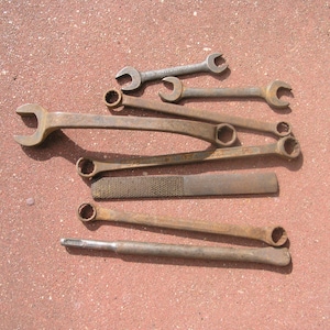 May include: A collection of vintage metal tools, including wrenches and a file, arranged on a textured, reddish-brown surface. The tools show signs of wear, with a rustic, aged appearance. The wrenches vary in size and style.
