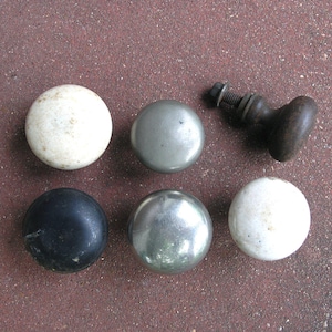 May include: A collection of six round cabinet knobs featuring various finishes. The set includes two white ceramic knobs, two metallic silver knobs, one matte black knob, and one dark brown wooden knob with an attached metal bolt and nut hardware.