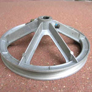 May include: A circular silver-colored metal pulley wheel featuring a central hub with a hollow core, five radiating support spokes, and a grooved outer rim. Two small metal set screws are visible near the central hub, one with a blue mark.