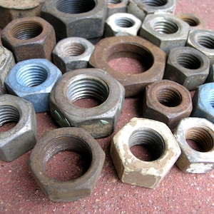 May include: A collection of assorted metal hex nuts in various sizes and finishes, including rusted, weathered, and galvanized steel. These industrial fasteners feature internal threading and hexagonal outer shapes, scattered together in a pile.