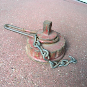 May include: A weathered, red metal cap with a chain and handle. The cap has a square top and a circular base with a ridged edge. The chain is attached to the cap and handle, and is made of silver metal.