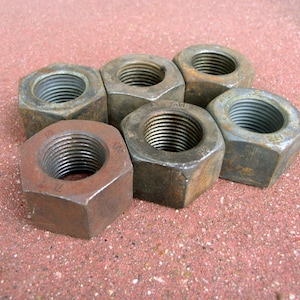 May include: Six rusty, hexagonal metal nuts with threaded interior holes are arranged on a reddish-brown surface. The nuts vary in color from dark gray to reddish-brown, showing signs of wear and age. The image is a close-up shot.