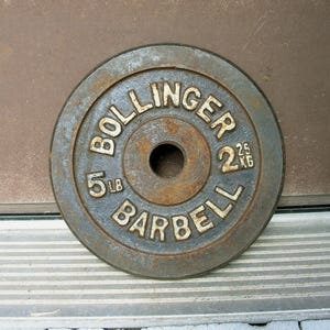 May include: A circular cast iron weight plate with a dark grey, weathered finish and visible rust. The text BOLLINGER BARBELL is embossed on the front, along with the weight markings 5 LB and 2.25 KG. It features a central hole for a barbell bar.