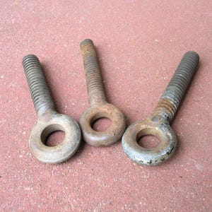 May include: Three heavy-duty metal eye bolts with threaded shanks and circular loops at the top. The hardware features a weathered, oxidized finish with visible signs of rust and surface wear, showing a metallic gray and brown color palette.